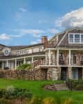 Large coastal house with stone foundation, wraparound porch, and manicured lawn under a blue sky.