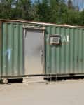 Green shipping container with a metal door, air conditioner, and chair outside, set on a dirt lot with trees in the background.