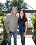 A couple standing in front of a small house, surrounded by a garden with red and white flowers.