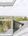Open balcony window with black railings overlooking a cityscape and a brick chimney with colorful pots.