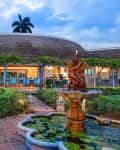 Tropical villa with a thatched roof, lush garden, and ornate fountain in the foreground.