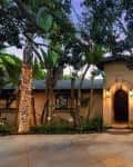 Single-story house with arched entrance, surrounded by palm trees and lush greenery, illuminated by warm exterior lights.