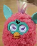 Pink furry toy with teal ears and eyes, orange beak, and purple tuft on a wooden table.