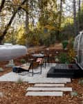 Airstream trailer in a wooded area with outdoor seating, table, and stepping stones surrounded by trees and foliage.