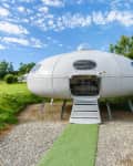 Futuristic UFO-shaped cabin with open door, set in a grassy field under a blue sky.