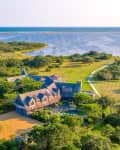 Aerial view of a large estate with a shingled house, surrounded by lush greenery, near a body of water.