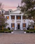 Colonial-style mansion with white columns, black shutters, and a gravel driveway, surrounded by trees and manicured hedges.