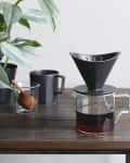 Coffee dripper on glass carafe, black mug, and plant on wooden table.