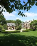 Large brick mansion with manicured lawn, surrounded by trees under a clear blue sky.