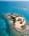 Small tropical island with blue-roofed buildings, palm trees, and a surrounding coral reef in clear turquoise water.
