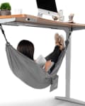 Person relaxing in a gray hammock under a wooden desk with a computer, plant, and stationery on top.