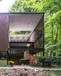 Modern two-story house with wooden facade, large windows, and open balcony surrounded by lush green forest.