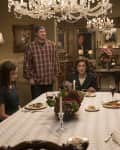 Elegant dining room with four people seated at a table, ornate chandelier, floral centerpiece, and classic paintings.