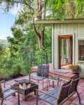 Patio with metal chairs and a fire pit, surrounded by lush trees and a small wooden cabin.