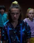 Five teenagers in colorful 80s outfits stand in a dimly lit room with brick walls.