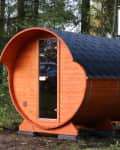 Wooden barrel sauna with a black shingled roof in a forest setting.