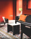 Modern lounge with black furniture, colorful cushions, and a patterned rug against orange walls.