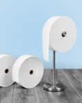 Three large toilet paper rolls, one on a metal stand, on a wooden floor against a blue wall.