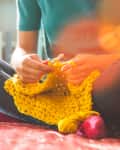 Person sitting cross-legged on a sofa, knitting with bright yellow yarn.