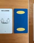 Three tarot cards on a wooden table: "The Llover," a blue card with yellow ovals, and "The World" with an IKEA illustration.
