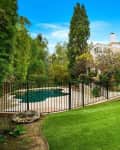 Two-story house with ivy-covered walls, fenced pool, lush garden, and manicured lawn under a clear blue sky.