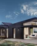 Modern beach house with solar panels, large windows, and outdoor seating on a wooden deck, surrounded by dunes.