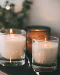 Three lit candles in glass holders on a table with blurred greenery and lights in the background.