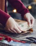 Hands wrapping a gift with brown paper on a wooden table, surrounded by scissors, ribbon, and holiday decor.