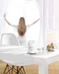 Woman opening curtains in bright room with white table, candle, and lamp.