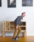 Child in gray hoodie and tan pants standing near a cardboard shelf, with framed photos on the wall above.