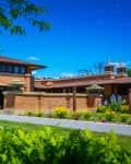 Brick building with large windows, surrounded by lush greenery and colorful flowers under a clear blue sky.