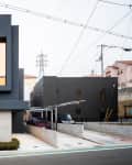 Modern narrow house with dark facade, large window, and minimal landscaping, situated between two other buildings.