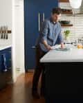 Man setting table in modern kitchen with blue cabinets, white countertops, and wooden floors.