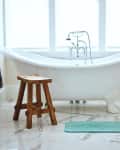 Freestanding bathtub with vintage faucet, wooden stool, and teal bath mat on marble floor.