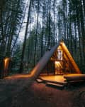 A-frame cabin with warm interior lighting surrounded by tall trees in a forest setting.