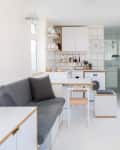 Modern white kitchen with gray sofa, small dining table, washing machine, and glass shower.