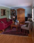 Living room with red sofa, patterned chairs, wooden armoire, and bookshelf, featuring a red rug and framed photos on walls.