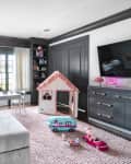 Playroom with gray sofa, toy house, dolls, and toy car on a pink rug, shelves with books and toys, and a wall-mounted TV.
