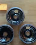 Three dirty stove burner drip pans on a wooden surface, labeled 1, 2, and 3 with sticky notes.
