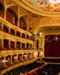 Ornate theater interior with red velvet curtains, gold balconies, and a painted ceiling, viewed from the audience seating area.
