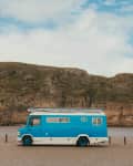 Blue camper van parked on a sandy beach with rocky cliffs in the background.