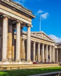 Neoclassical building with tall columns, people walking and sitting on the grass under a clear blue sky.