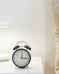 Alarm clock on a bedside table next to a modern lamp and striped pillows.