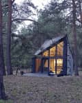 A-frame cabin with large glass windows in a forest, warm interior lighting visible, surrounded by tall pine trees.