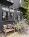 Dark gray building facade with large windows, wooden bench, and purple flowering plants on cobblestone street.