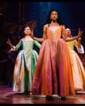 Performers in period costumes on stage, with a woman in an orange dress leading, surrounded by others in colorful gowns.