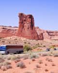 Tour bus and cars on a desert road with red rock formations under a clear blue sky.