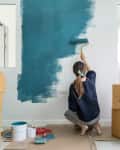 Woman painting a wall teal in a room with cardboard boxes and a potted plant.