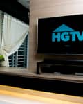 Modern living room with large TV displaying HGTV logo, wooden paneling, and decorative plants on a shelf.