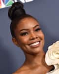 Woman smiling on red carpet in white ruffled dress, hair in a bun, with NBC and #AGT logos in the background.